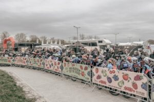 Crowd of cyclists wearing helmets lined up behind a fruit-themed barrier at an outdoor event, with campers in the background under a cloudy sky.