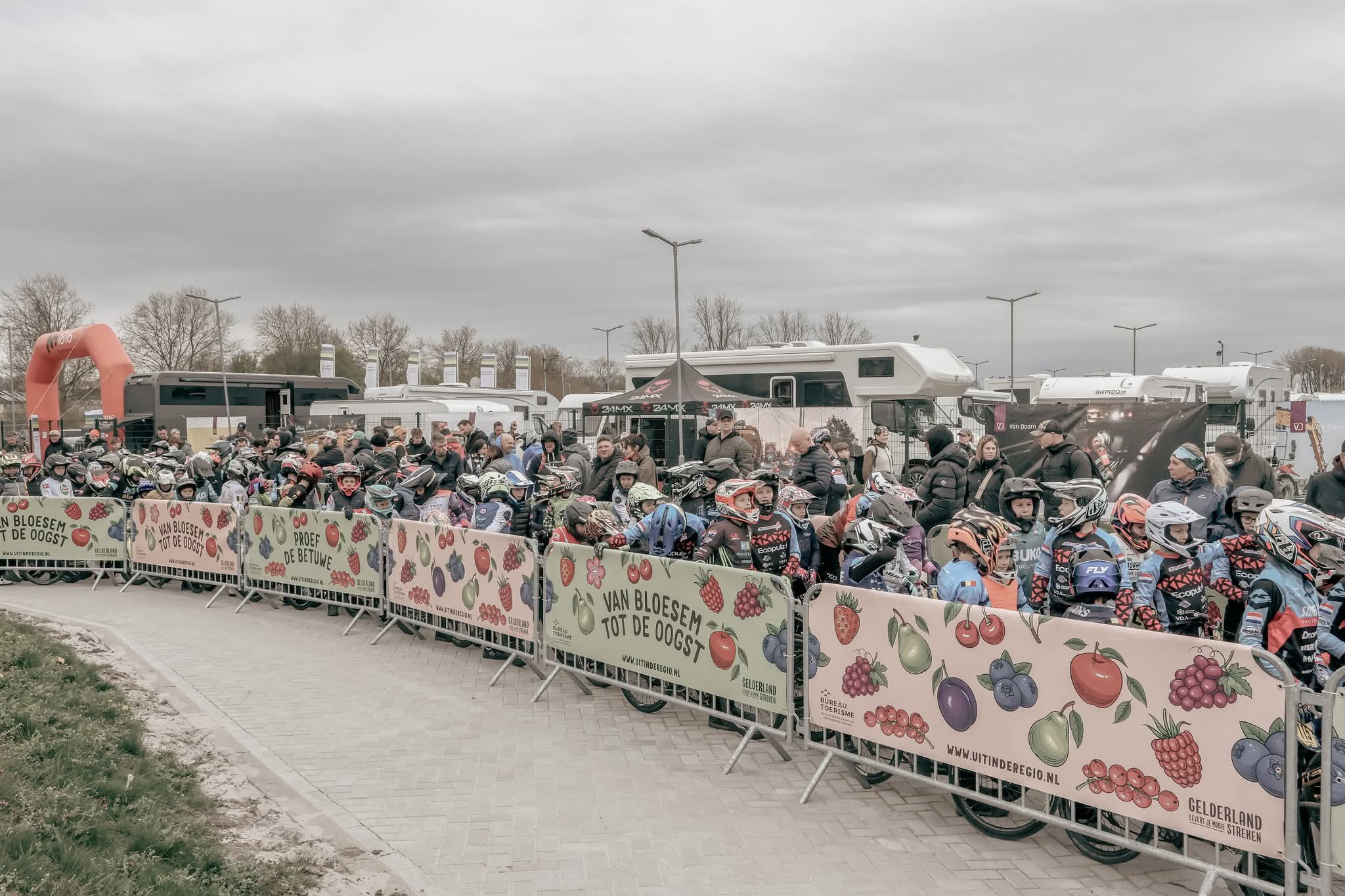 Crowd of cyclists wearing helmets lined up behind a fruit-themed barrier at an outdoor event, with campers in the background under a cloudy sky.