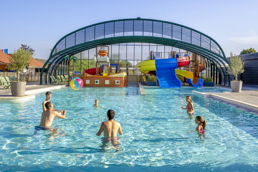 Outdoor pool with colorful water slides under a curved glass canopy; people playing in the water on a sunny day.