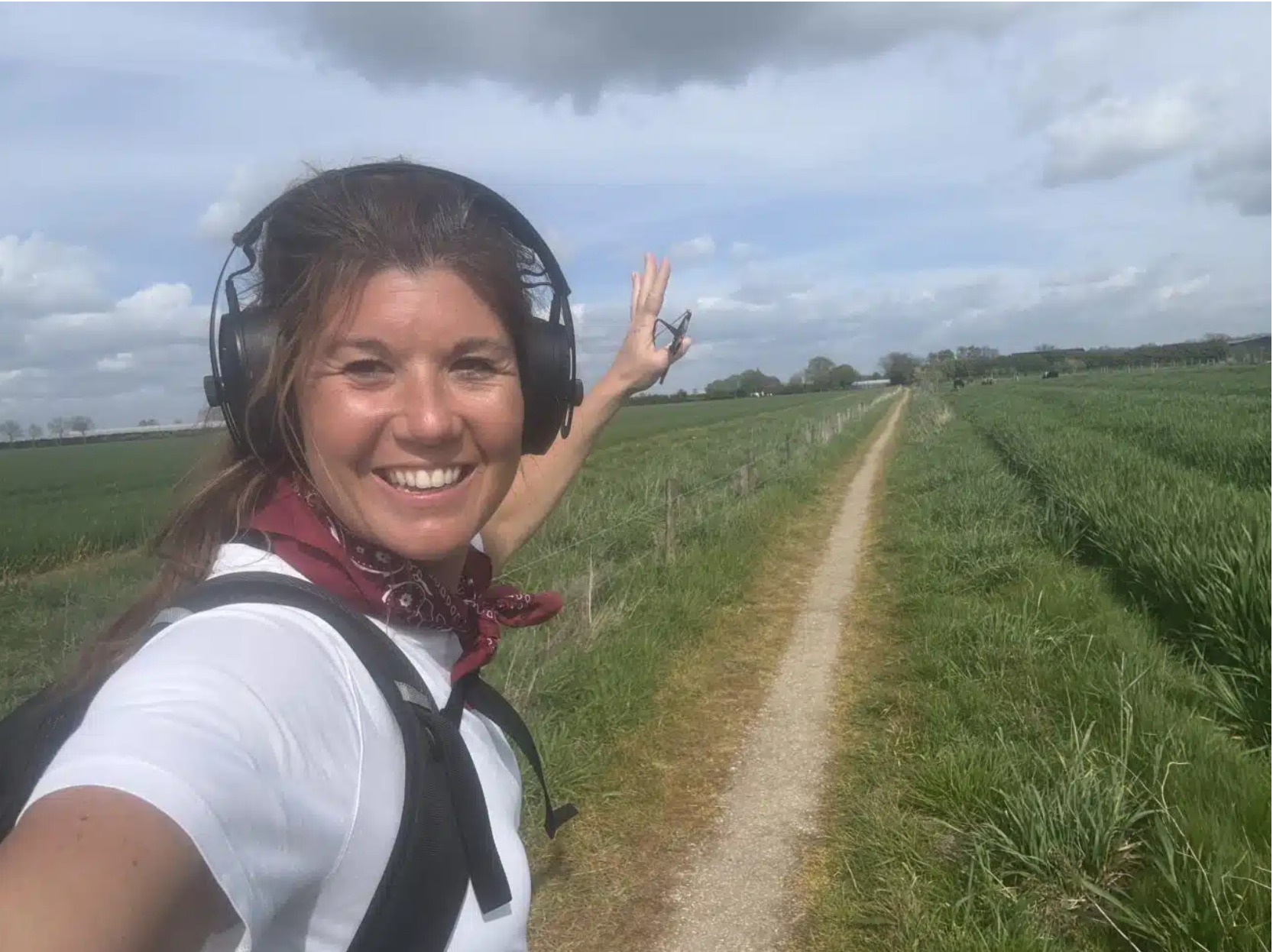 Smiling woman wearing over-ear headphones and a backpack, taking a selfie on a rural dirt path with green fields on either side and a cloudy sky above.