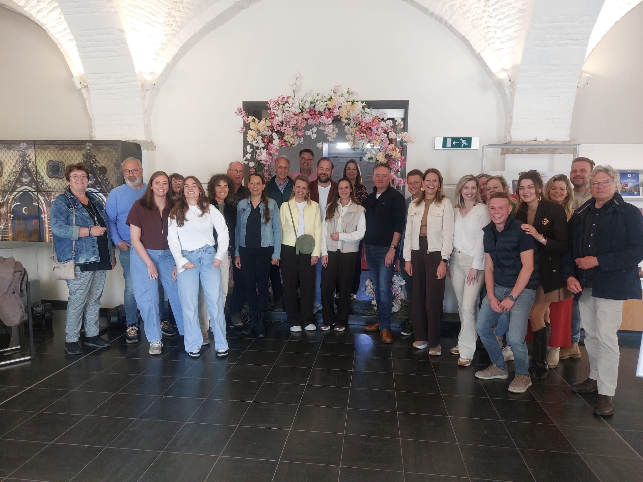 Group of about 25 adults posing for a photo indoors in front of a pink floral arch beneath white arched ceiling.