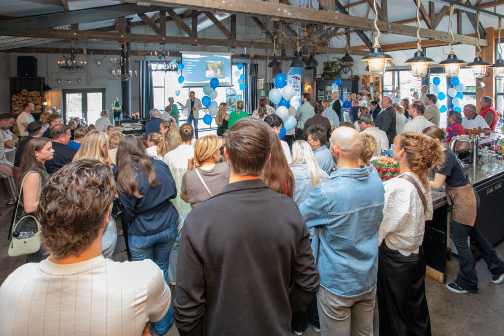 Large crowd gathered in a rustic indoor venue for a presentation, with a speaker on stage and blue and white balloons.