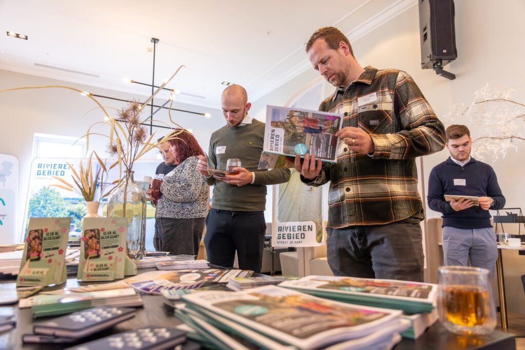 Four people stand around a table studded with brochures, magazines and a glass of drink, reading and browsing at an indoor event.