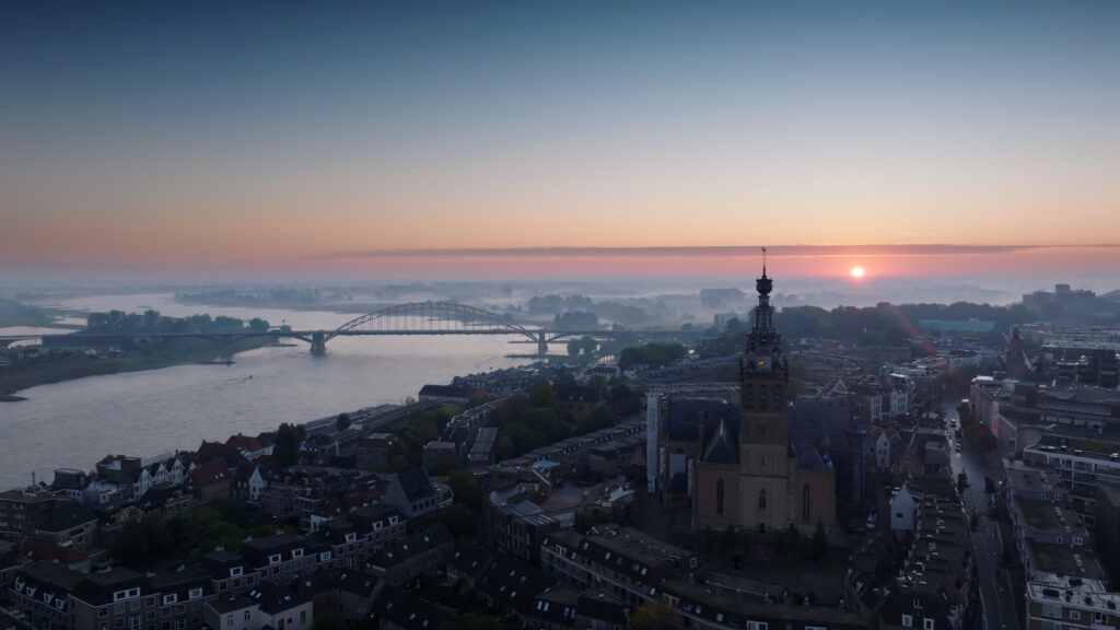 Aerial view of a city at sunset with a river, an arched bridge, and a tall historic church spire in the foreground.