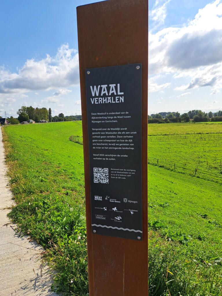 Rusty brown metal post with a black plaque reading 'Waal Verhalen' beside a green field under a blue sky; plaque includes small text and a QR code.