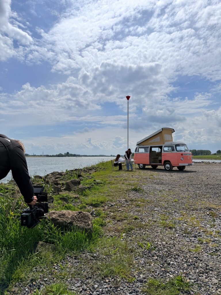 Retro pink VW camper with pop-up roof parked on a rocky lakeside; two people set up a table while a photographer films nearby.
