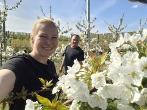 Smiling woman taking a selfie in a blossoming orchard with white flowers on the trees and a man in the background.
