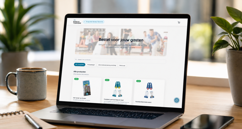 webshop - Stichting Bureau Toerisme Laptop on a wooden desk showing a Dutch travel site with a search bar and product cards; a mug sits to the left and plants surround the scene.