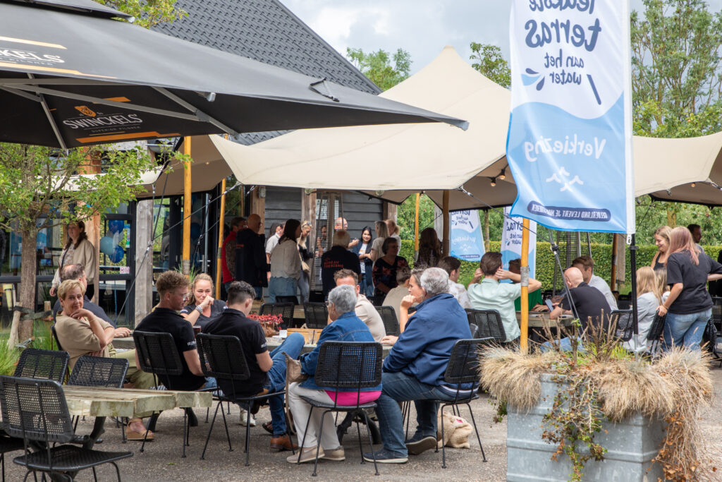 Outdoor daytime gathering with people seated at black wicker chairs under large beige tents and umbrellas.
