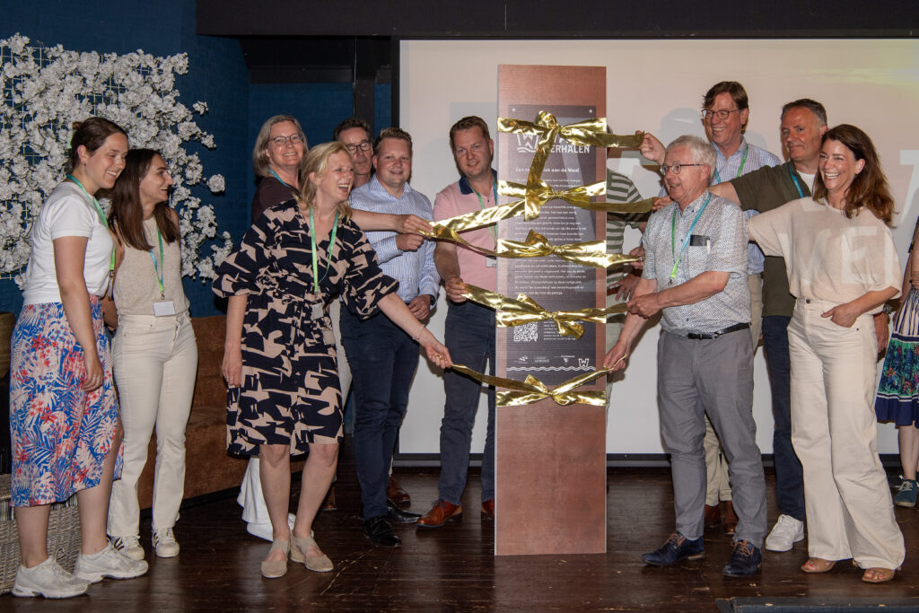 Group of people smiling during a ribbon-cutting/launch event around a tall poster wrapped with gold ribbons inside a room with a wooden floor.