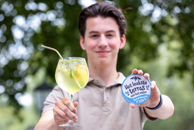 Young man outdoors presenting a lime cocktail and a circular award reading 'Het leukste terras aan het water'.