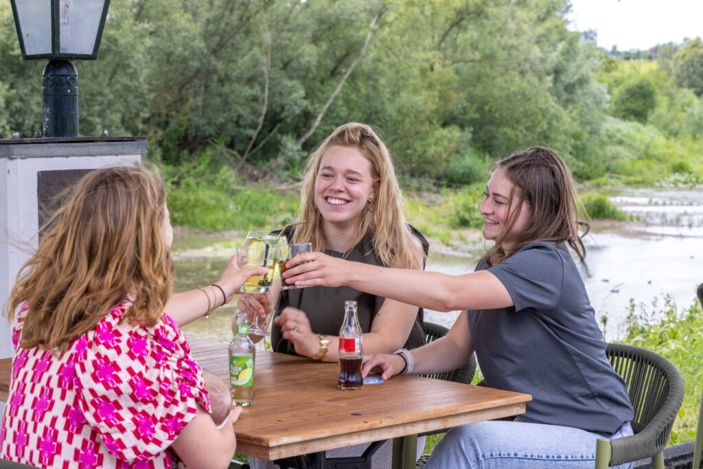 Three women at an outdoor riverside table toasting with glasses, smiling as drinks raised toward the center.