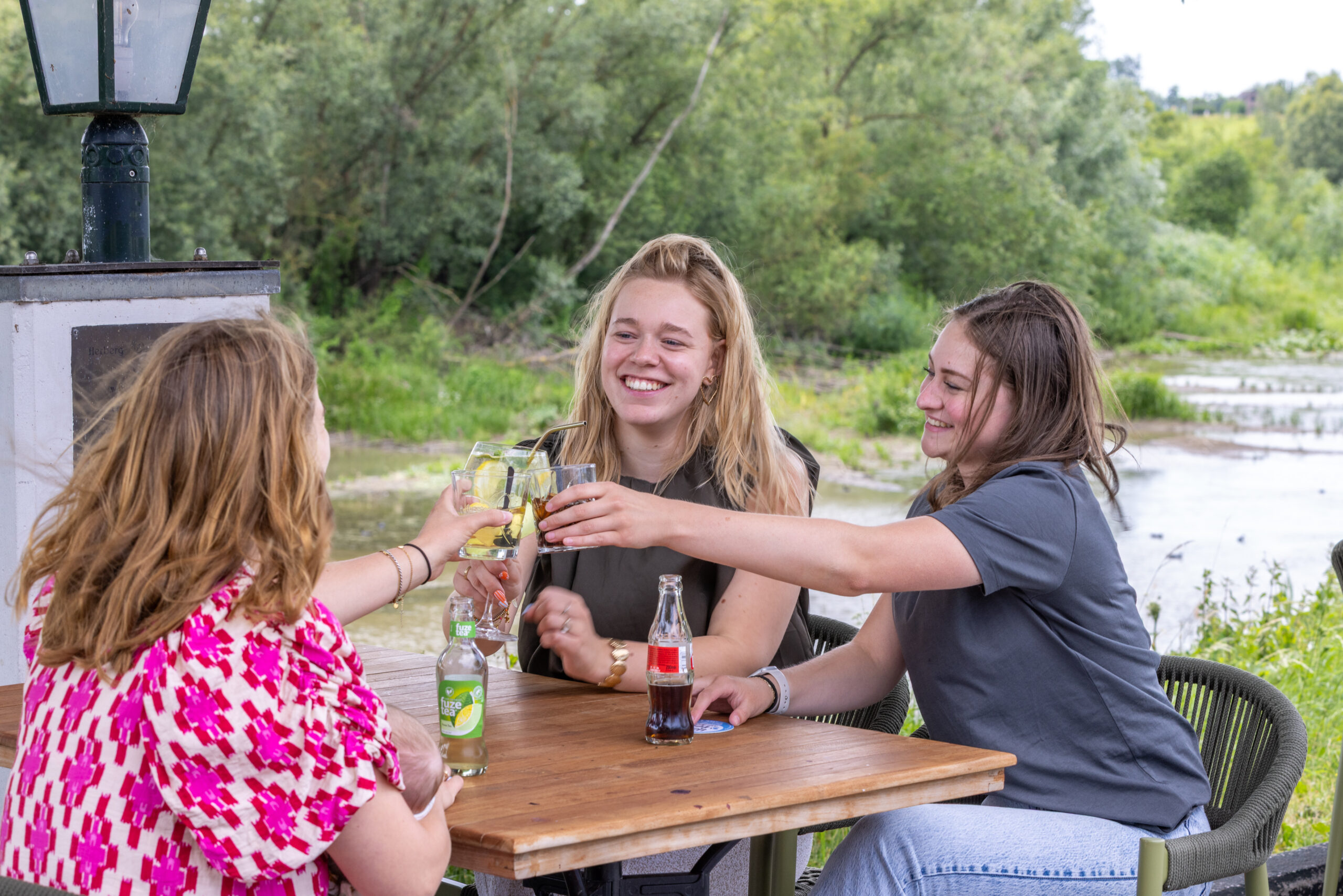 Three women at an outdoor riverside table toasting with glasses, smiling as drinks raised toward the center.