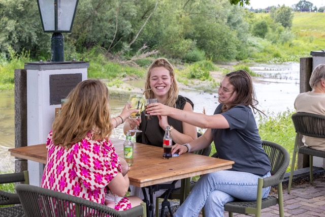 Three women toast with drinks at a wooden table outdoors by a river, smiling.