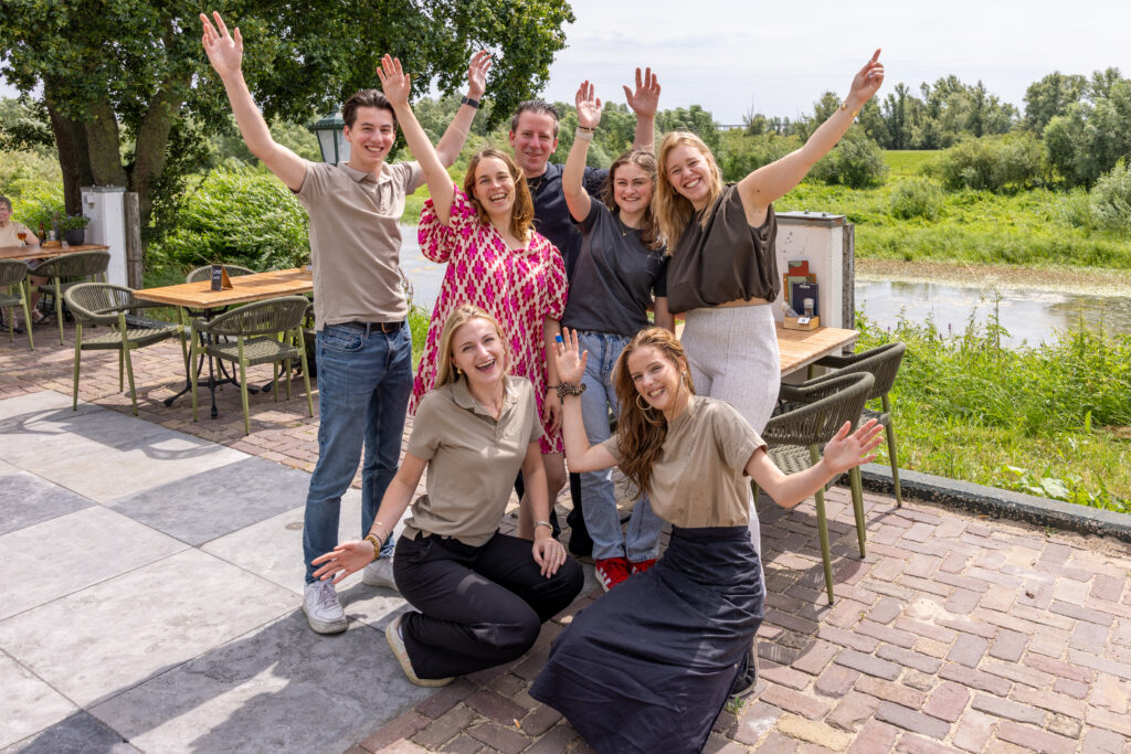 Group of smiling people outdoors posing with raised arms, near a river and cafe tables on a sunny day.
