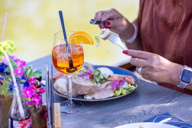 Aperol Spritz with orange slice on a sunny outdoor table; plate of fish, bread, and salad nearby as a person cuts food with a fork and knife.