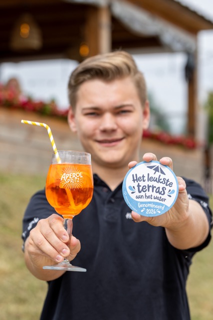 Young man outdoors holding an orange drink with a striped straw in one hand and a round award badge reading 'Het leukste terras aan het water' in the other.