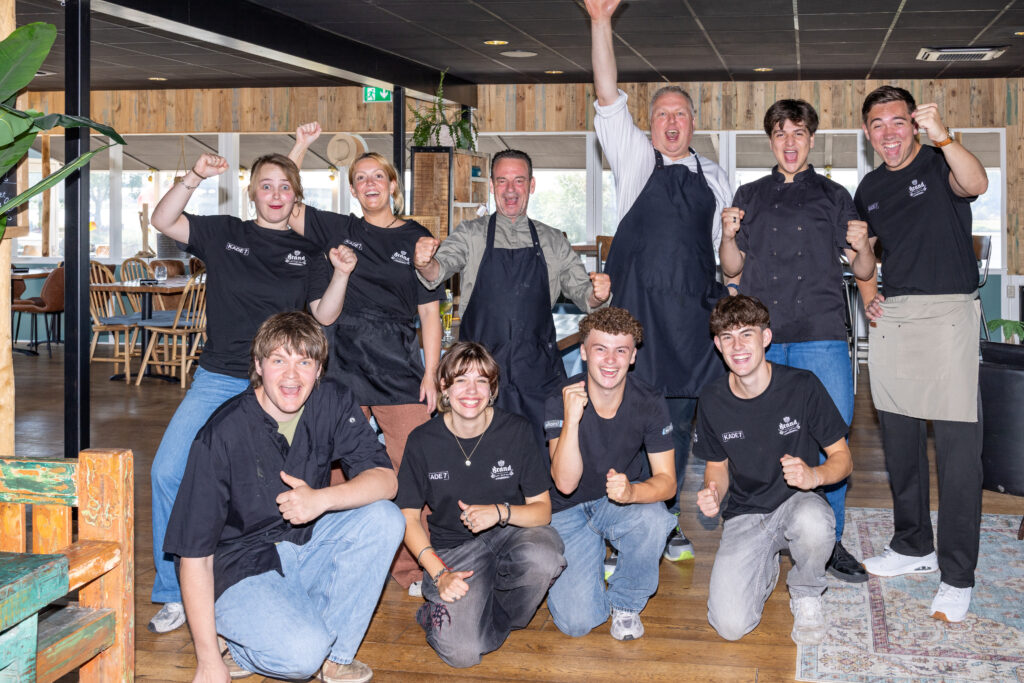 Group of smiling staff in matching black shirts and aprons posing with fists raised indoors at a restaurant/bar setting habitually used for team photos.