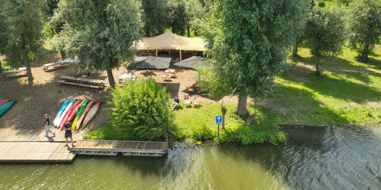 Riverside area with a large beige tent, picnic tables, and colorful kayaks lined on a wooden dock by the water.