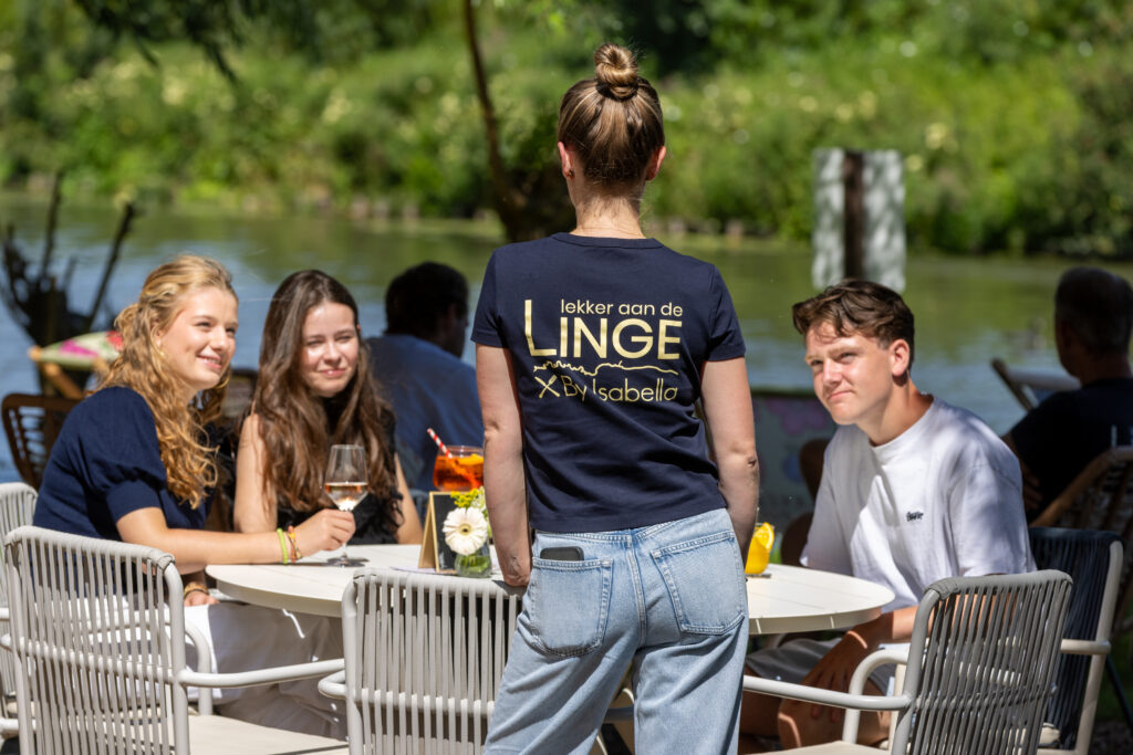 A group of friends seated at a white table outdoors by a lake, with a server standing with back to camera wearing a navy T-shirt.