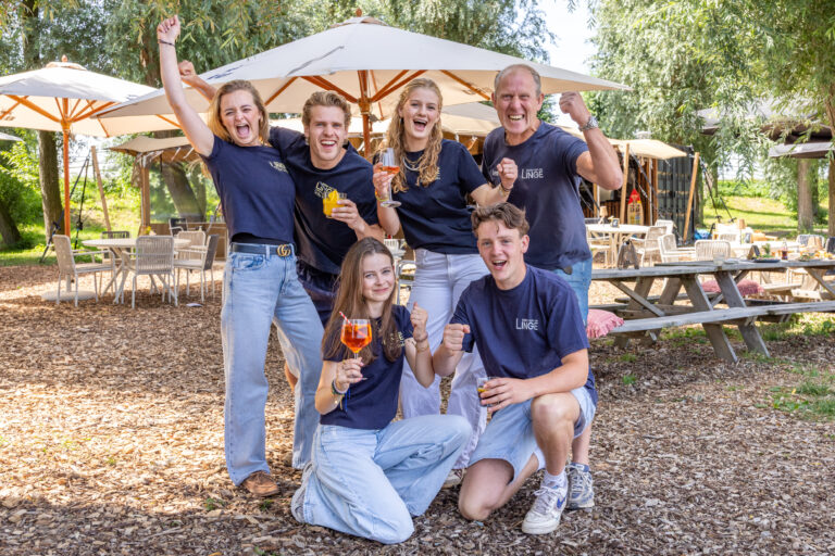 Six friends cheer and pose for a photo outdoors, each holding a drink under cafe-style umbrellas and trees in a sunny setting.