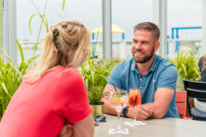 Two people chat at an outdoor cafe table with drinks, greenery around, man in blue shirt smiling at a woman in red.