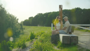 Couple sitting and chatting on a wooden park bench beside a grassy path on a sunny day, with trees in the background.