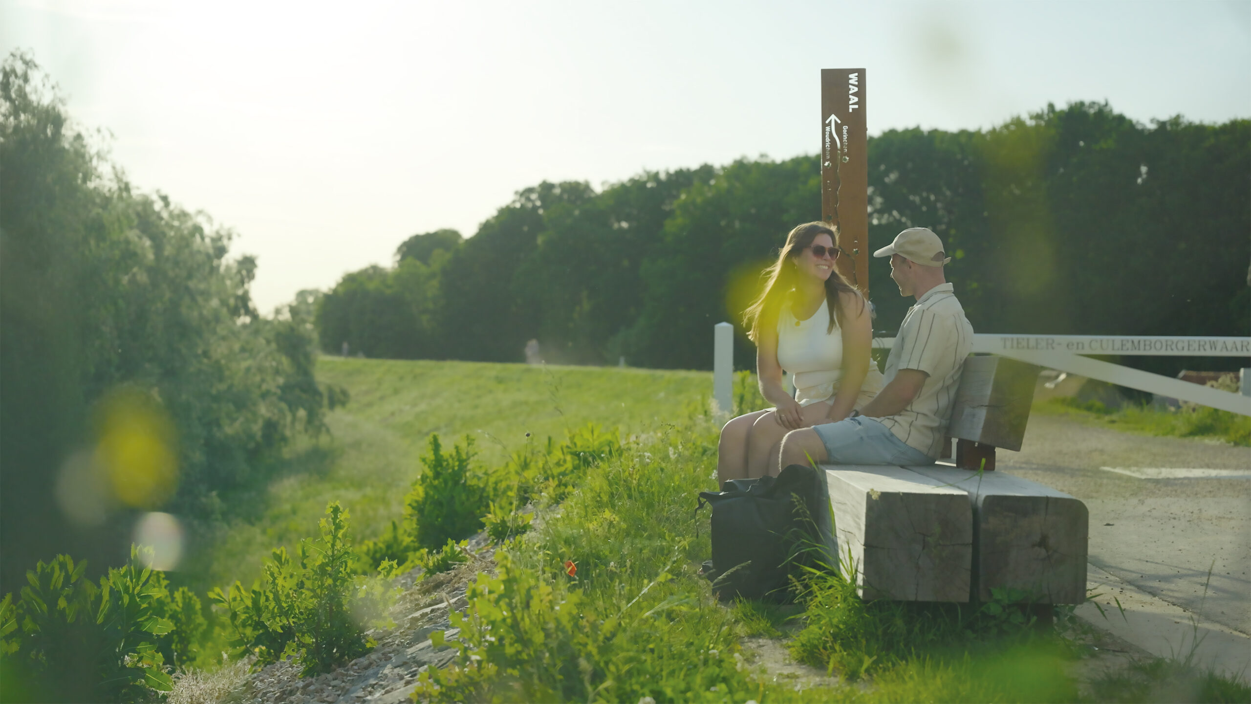 SK Waalzuil foto - Neerijnen - Zuil_001 - Waalverhalen - Stichting Bureau Toerisme - Stichting Bureau Toerisme Couple sitting and chatting on a wooden park bench beside a grassy path on a sunny day, with trees in the background.