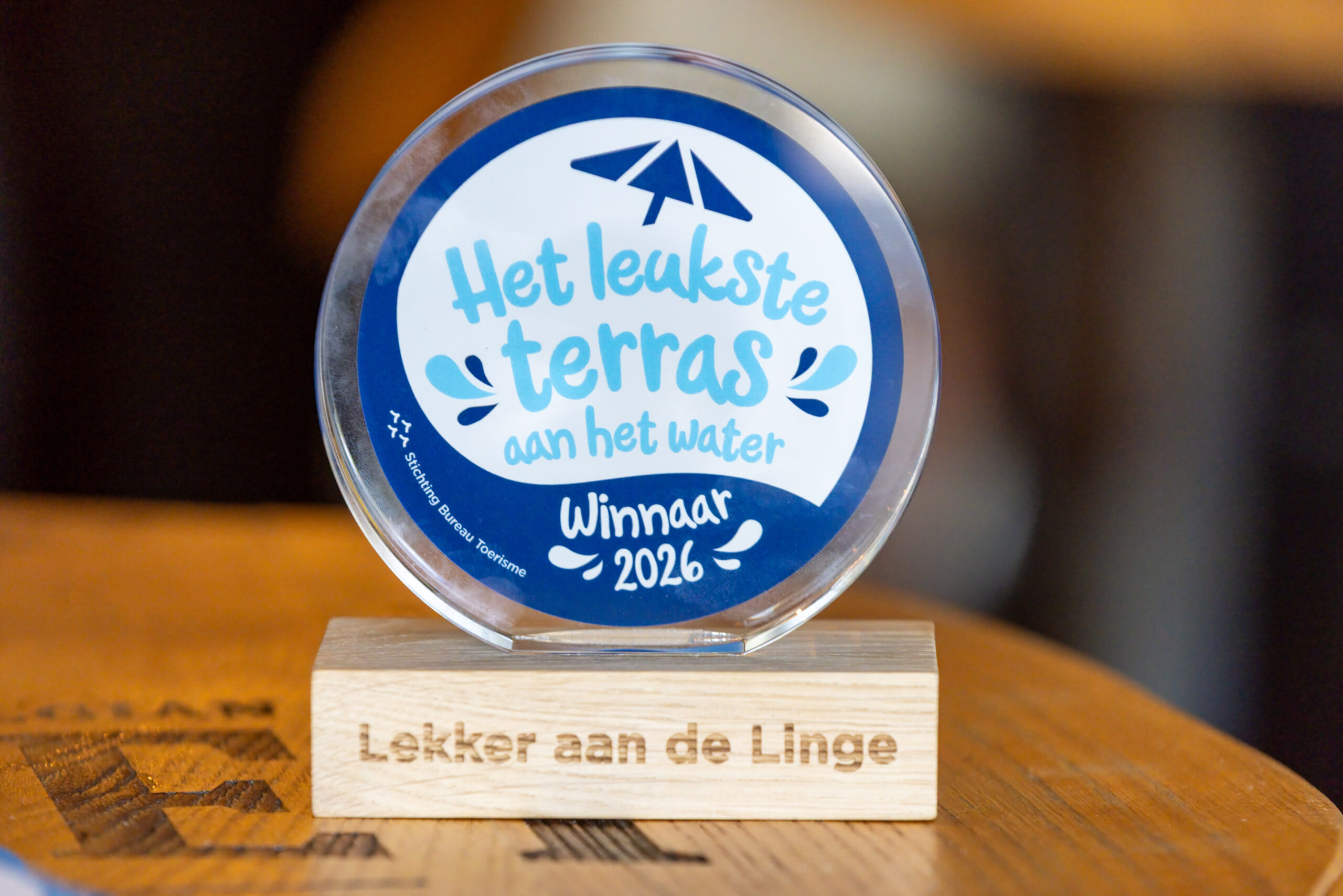 Round glass award with a blue ring, displaying 'Het leukste terras aan het water' and 'Winnaar 2026' on a wooden stand.