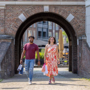 Two friends walk with shopping bags under a brick archway on a sunny day, smiling at the camera.
