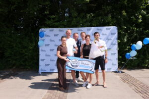 Six people pose outdoors with a first‑place banner and bouquet, in front of a logo backdrop and blue balloons.