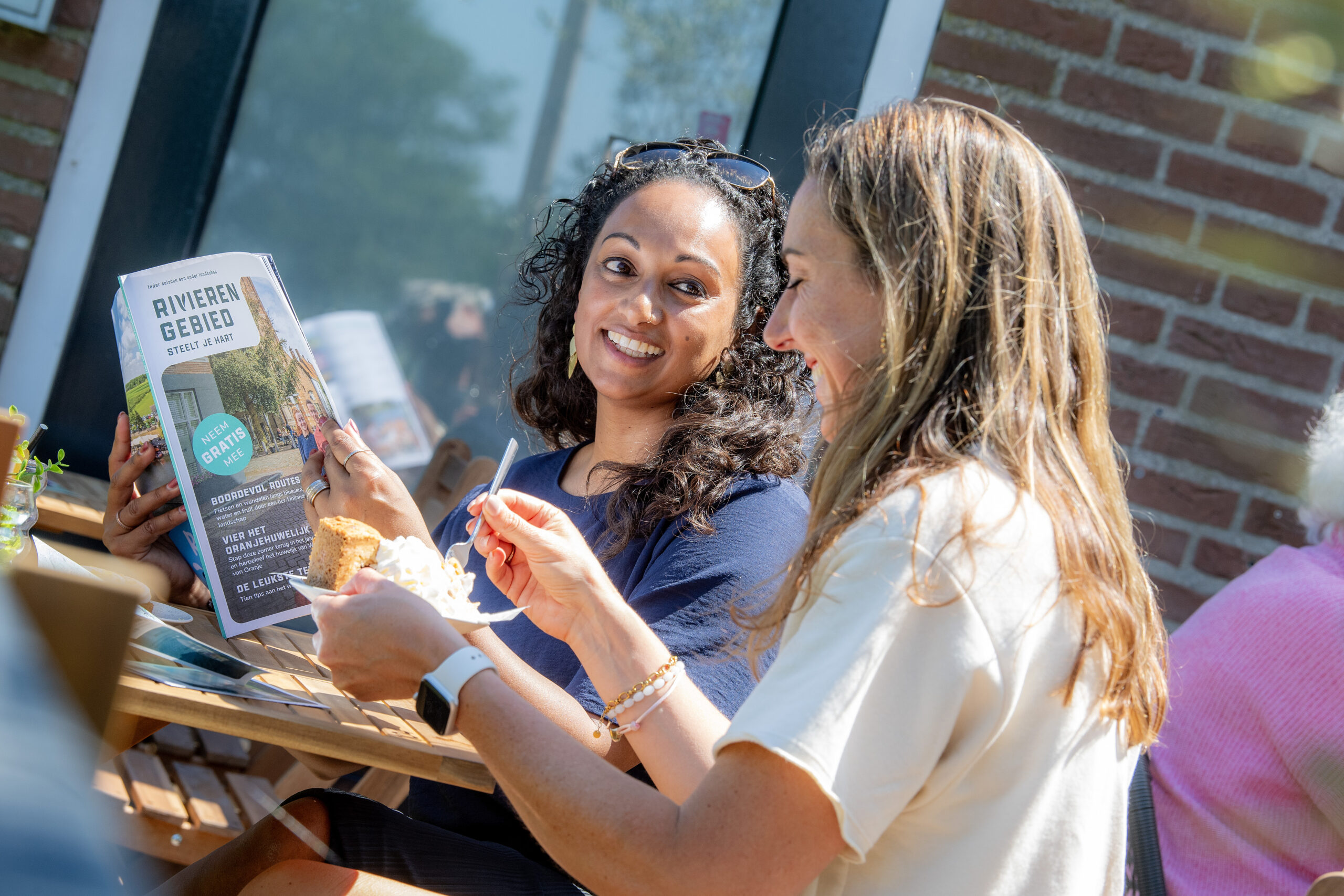 Two women at an outdoor café share a smile while one shows a brochure titled 'Rivieren Gebied' and they enjoy dessert.