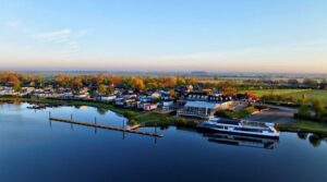 A tranquil riverside town with a marina, houses, trees, and a large white passenger boat docked by calm water at sunset.
